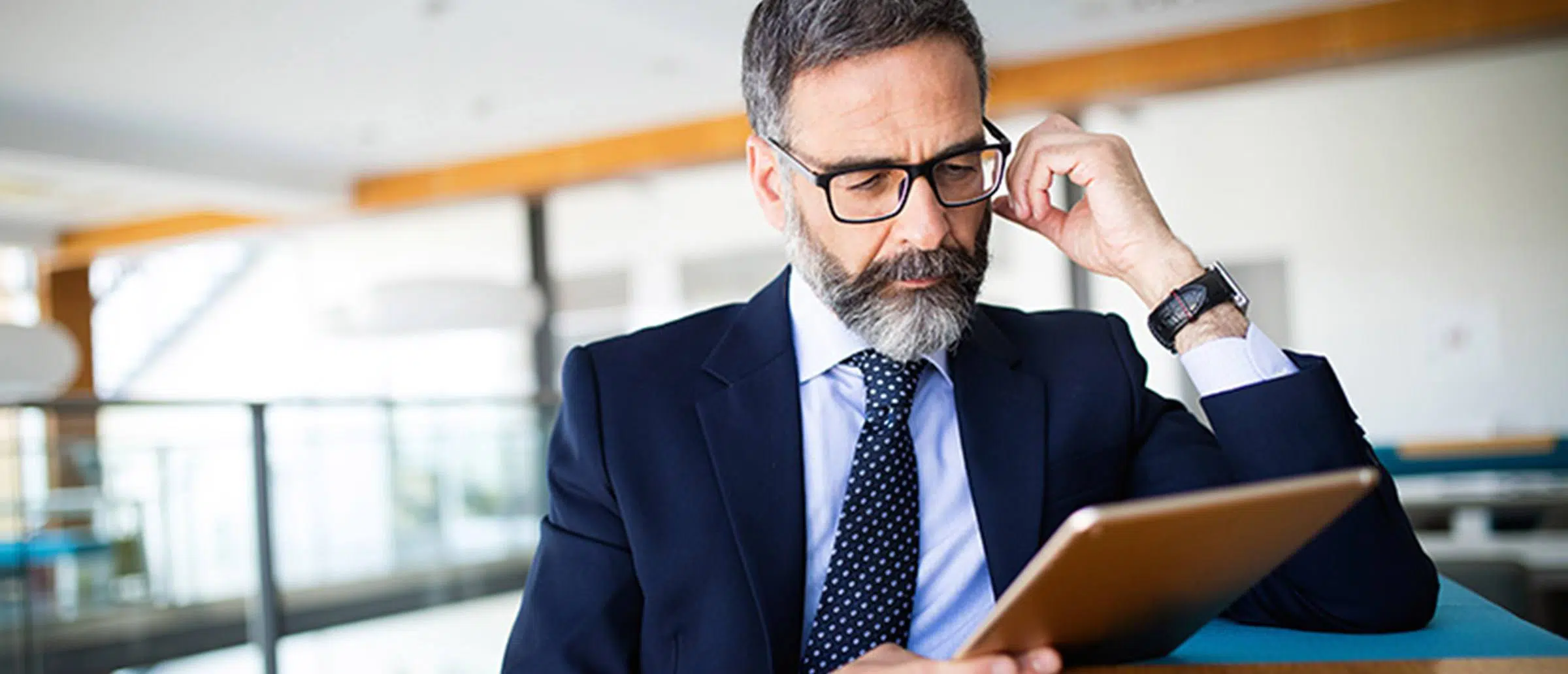 A middle-aged, bearded businessman with glasses in a suit and tie reading a tablet device