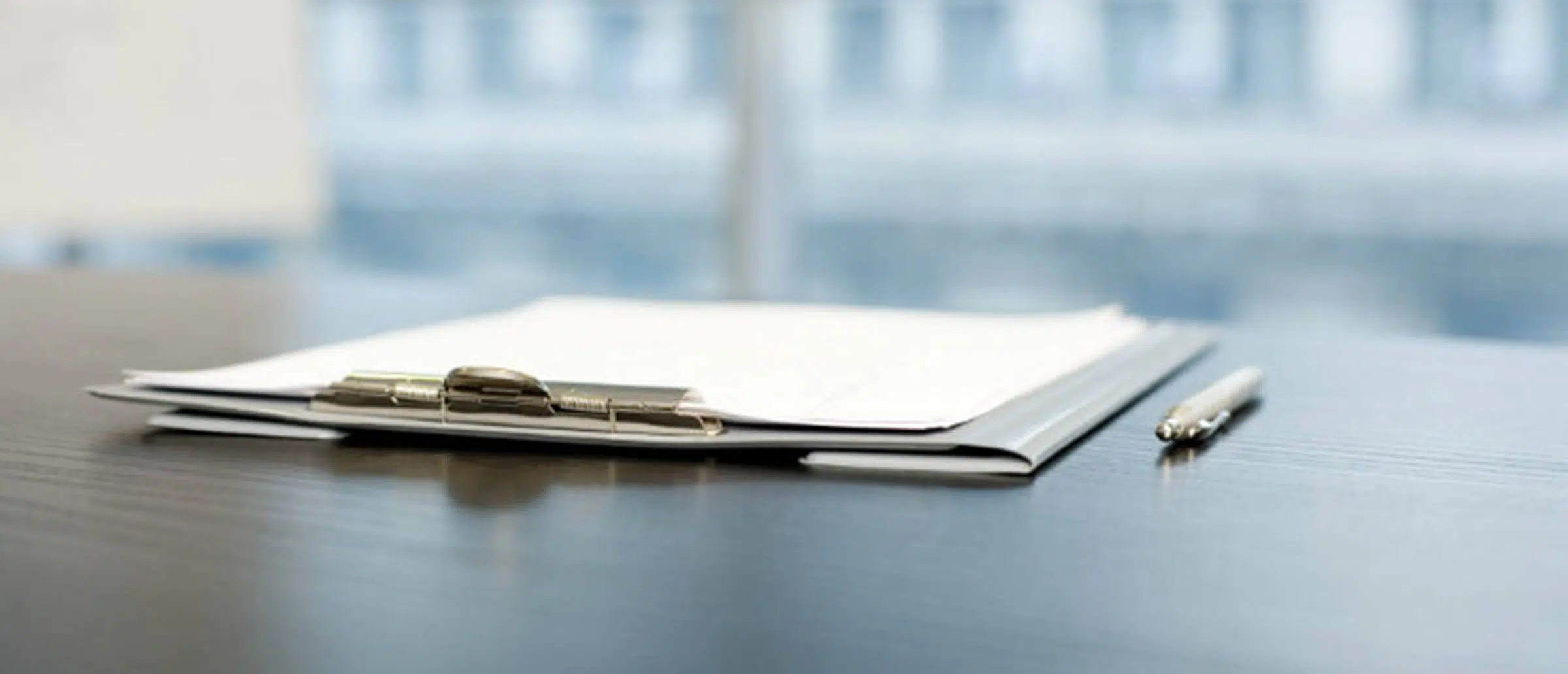 Pen and clipboard with paper laid out on a grey table with a blurred glass window in the background
